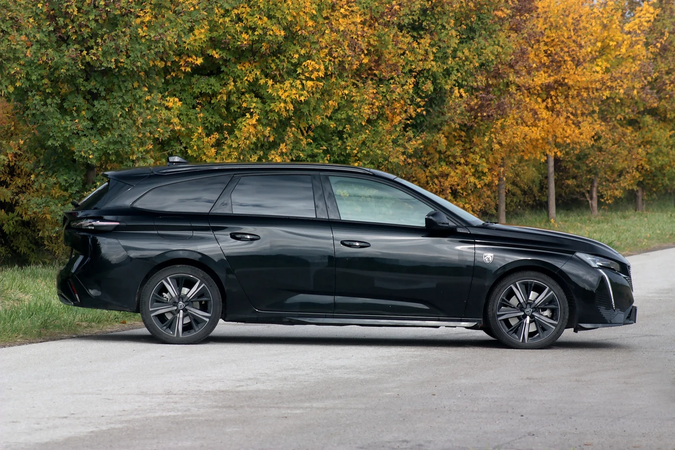 Czarny samochód osobowy typu kombi zaparkowany na poboczu drogi, w tle drzewa w jesiennych barwach żółci i pomarańczy.