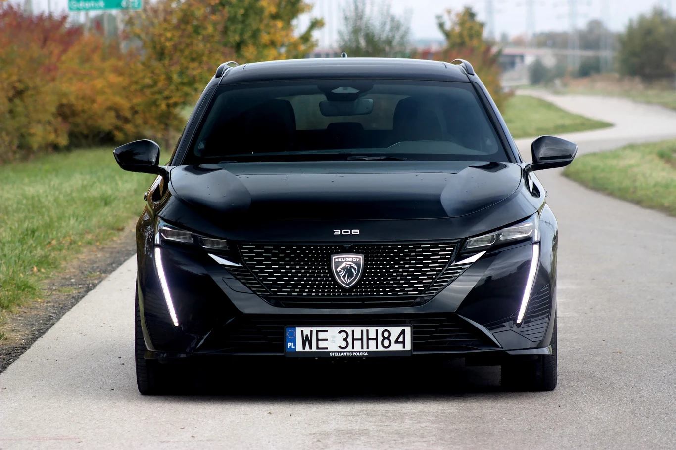 Czarny samochód Peugeot 308 stoi na wąskiej asfaltowej drodze otoczonej zielenią i kolorowymi drzewami, na pierwszym planie widoczny jest charakterystyczny grill i światła LED.