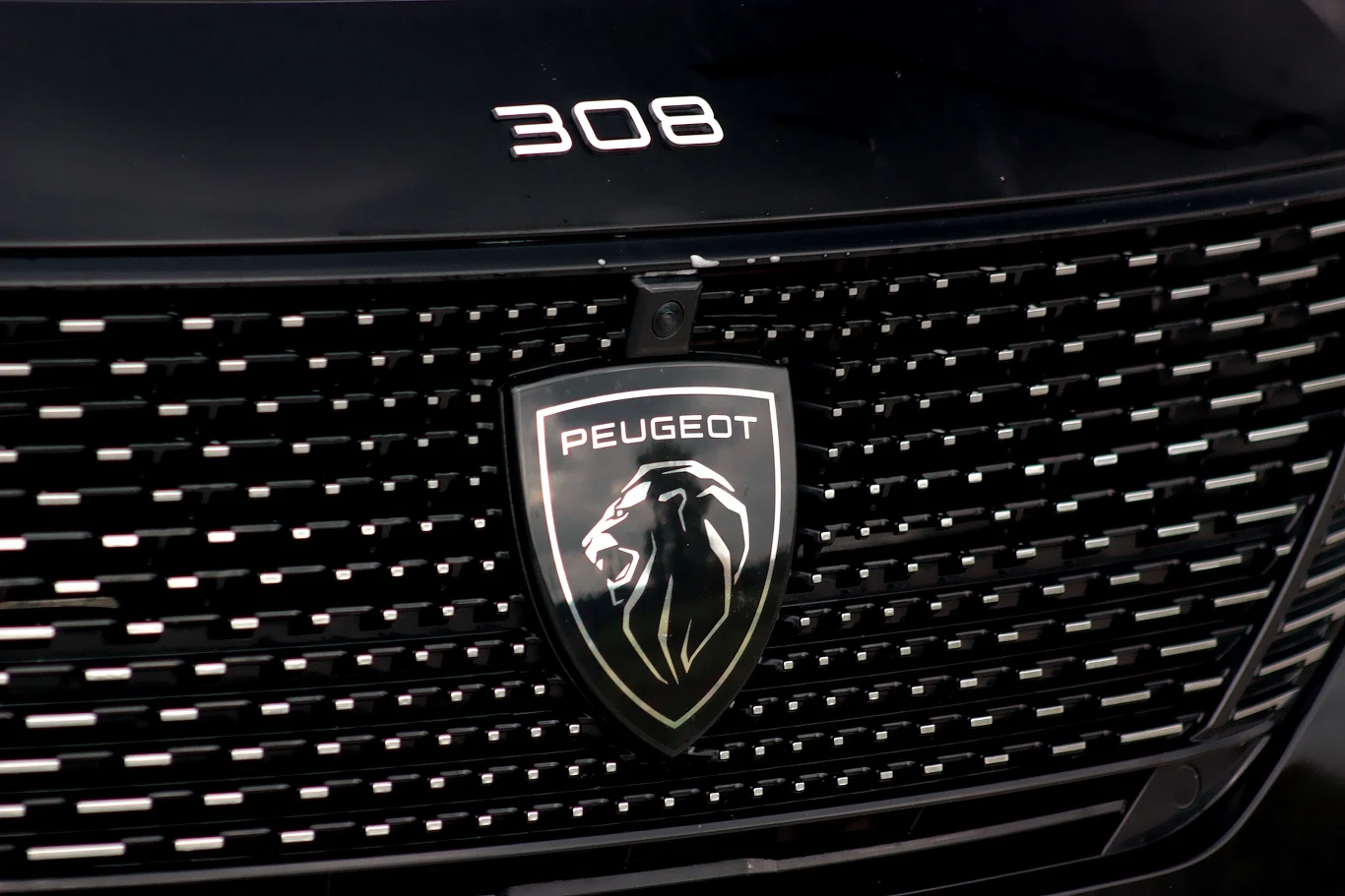 Maska samochodu z widocznym emblematem marki Peugeot oraz numerem modelu 308, na tle czarnej osłony chłodnicy z charakterystycznym wzorem.
