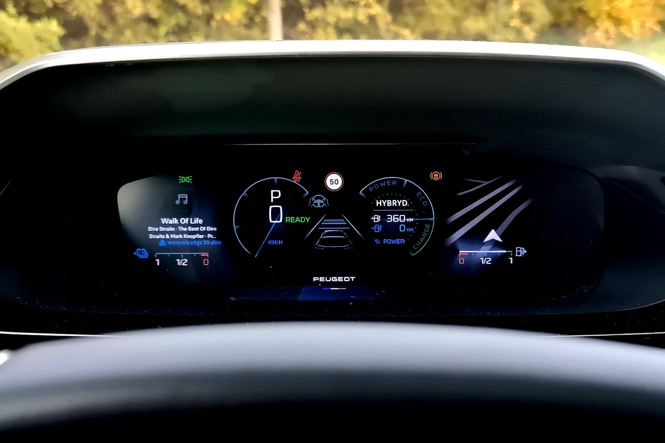 Cyfrowy panel wskaźników nowoczesnego samochodu z wyraźnie widocznymi informacjami o prędkości, stanie akumulatora hybrydowego, wskazaniami systemu nawigacji oraz aktywnym odtwarzaczem muzyki. Otoczenie panelu jest dobrze oświetlone, przez co widoczne ...