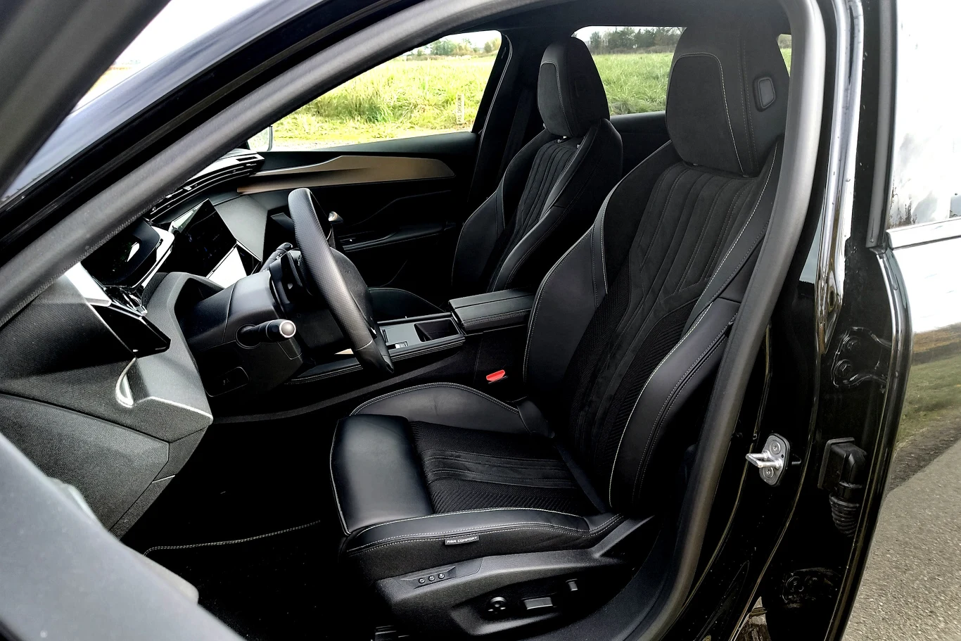 Nowoczesne, przestronne wnętrze samochodu z dwoma przednimi, czarnymi fotelami, widoczną kierownicą oraz panelem środkowym. Jasne światło wpada przez boczne okno, a za samochodem rozciąga się zielony krajobraz.