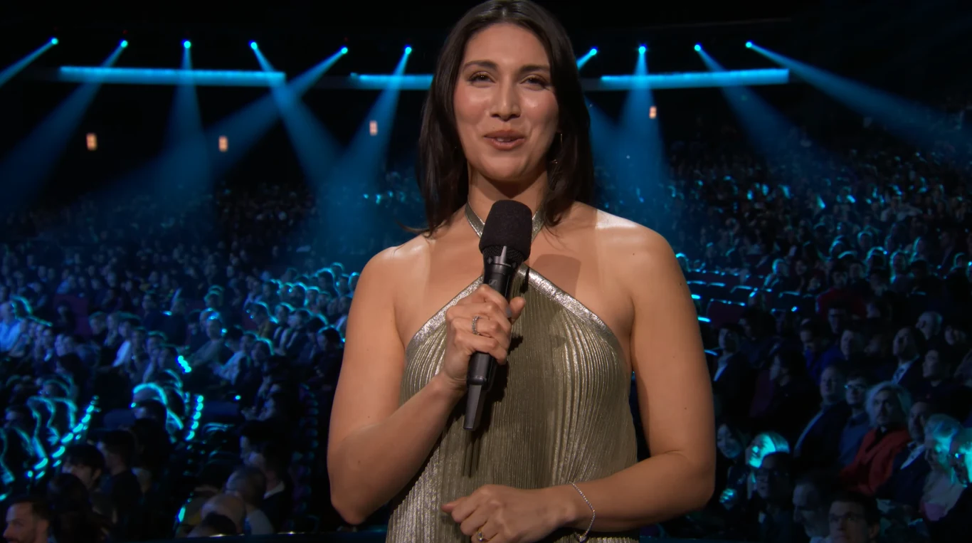 The Game Awards 2025 Uśmiechająca się kobieta w eleganckiej sukni trzyma mikrofon na tle dużej publiczności podczas uroczystego wydarzenia w hali oświetlonej niebieskawymi reflektorami.
