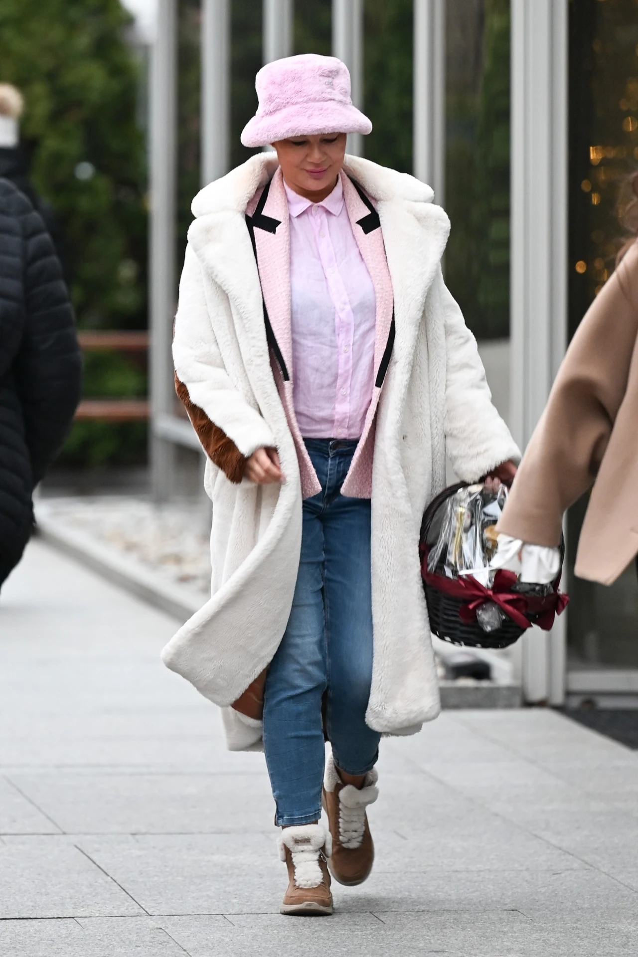 Edyta Górniak przyłapana przed siedzibą Polsatu Kobieta ubrana w biały, futrzany płaszcz, jasnoróżową koszulę i różowy kapelusz spaceruje chodnikiem miejskim, trzymając w jednej ręce torebkę i zakupy. W tle widoczne są inne osoby oraz nowoczesna zabudowa.