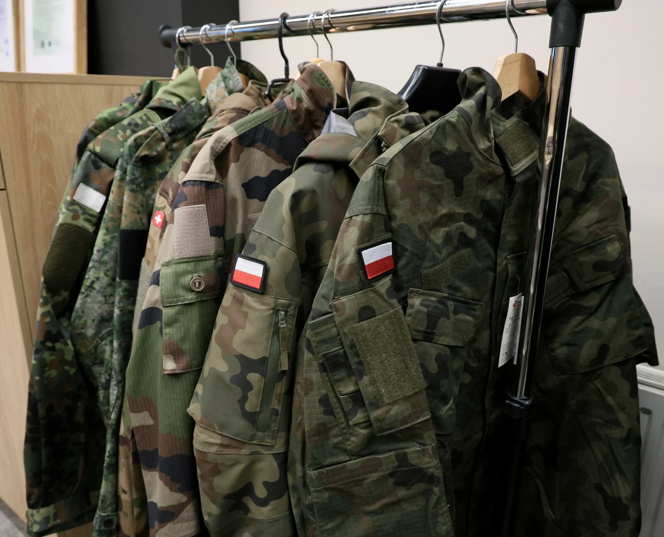 Promocje w sklepie Agencji Mienia Wojskowego na nietypowe produkty Wiszące na wieszaku polskie mundury wojskowe w różnych wzorach kamuflażu, wyraźnie widoczne naszywki z flagą Polski na każdym z nich.