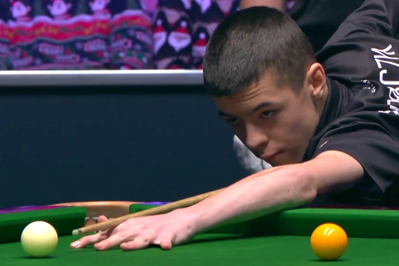 Młody mężczyzna w skupieniu wykonuje uderzenie kijem bilardowym nad zielonym stołem do snookera, koncentrując się na białej bili. W tle znajduje się ciemny wzorzysty panel i część publiczności.