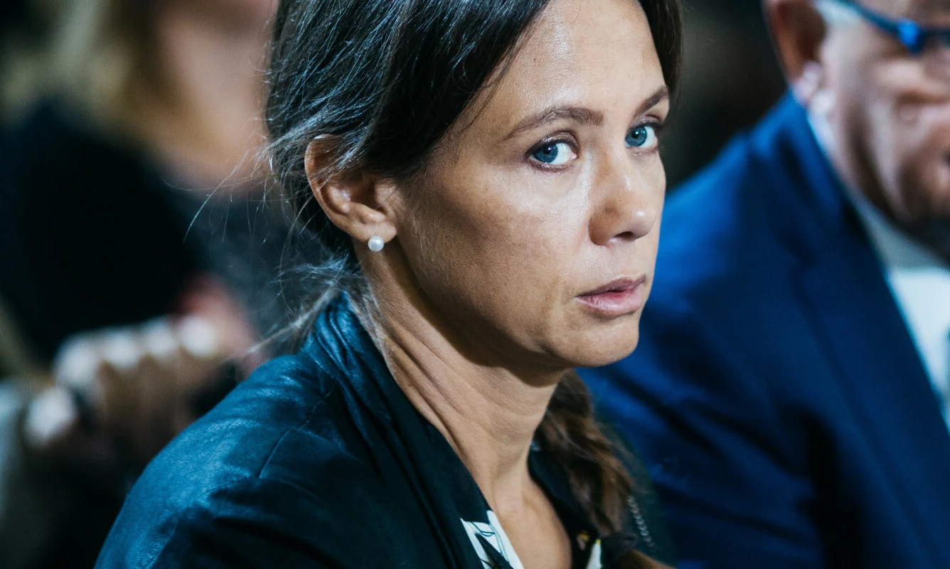 Kinga Rusin Kobieta w ciemnej marynarce z perłowymi kolczykami patrzy w stronę aparatu, obok niej siedzi mężczyzna w okularach i niebieskim garniturze.