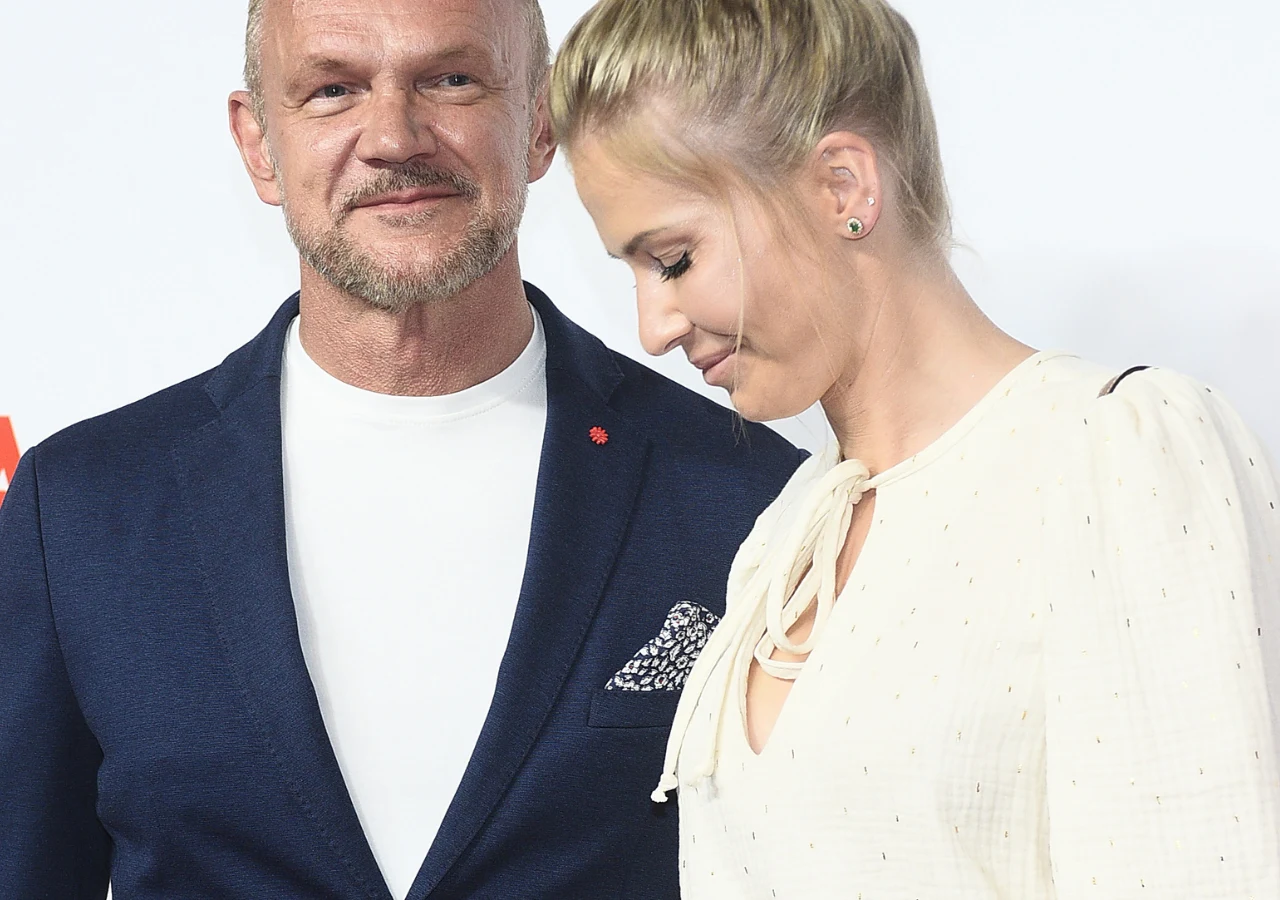 Edyta Pazura, Cezary Pazura Dwie osoby stoją obok siebie na jasnym tle, mężczyzna ubrany w granatową marynarkę z białą koszulką uśmiecha się, kobieta w jasnej sukience z upiętymi blond włosami patrzy w dół i delikatnie się uśmiecha.
