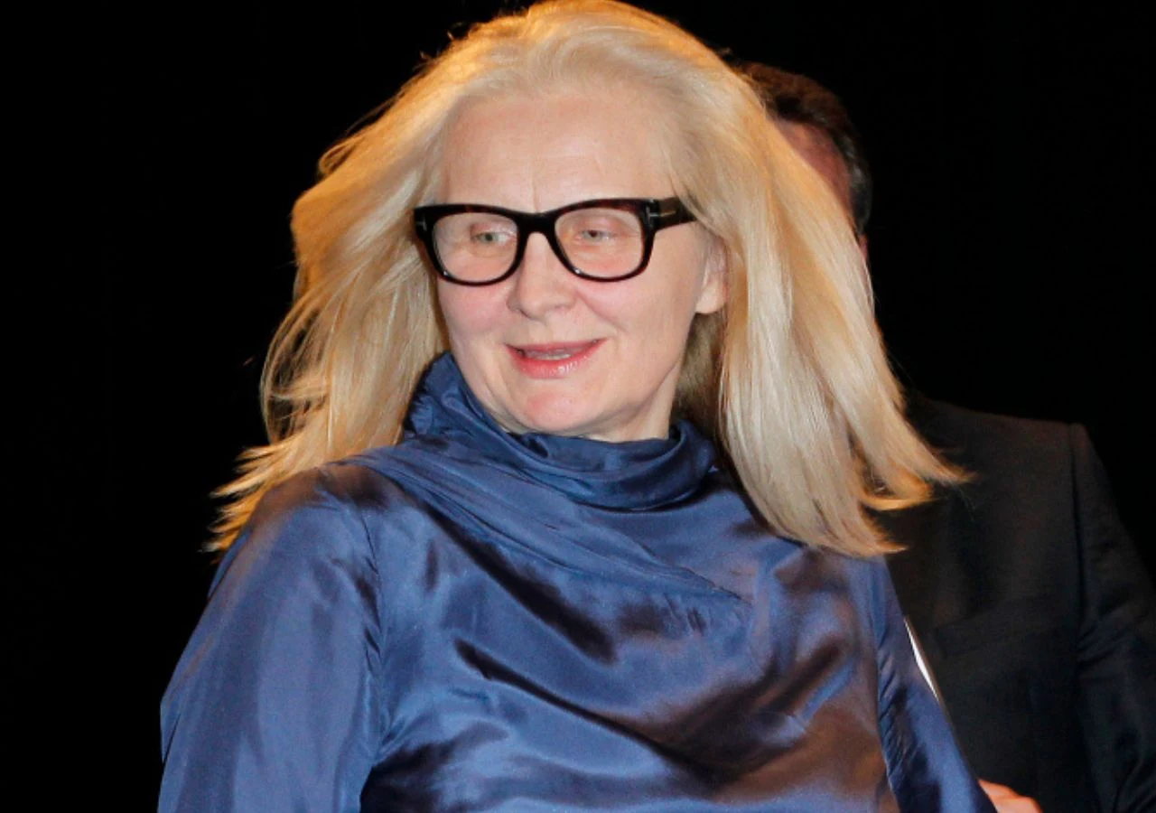 Magda Umer w 2011 roku Kobieta w średnim wieku z długimi blond włosami i dużymi czarnymi okularami, ubrana w satynową granatową bluzkę, stoi na ciemnym tle. Uśmiecha się i wygląda na zadowoloną.