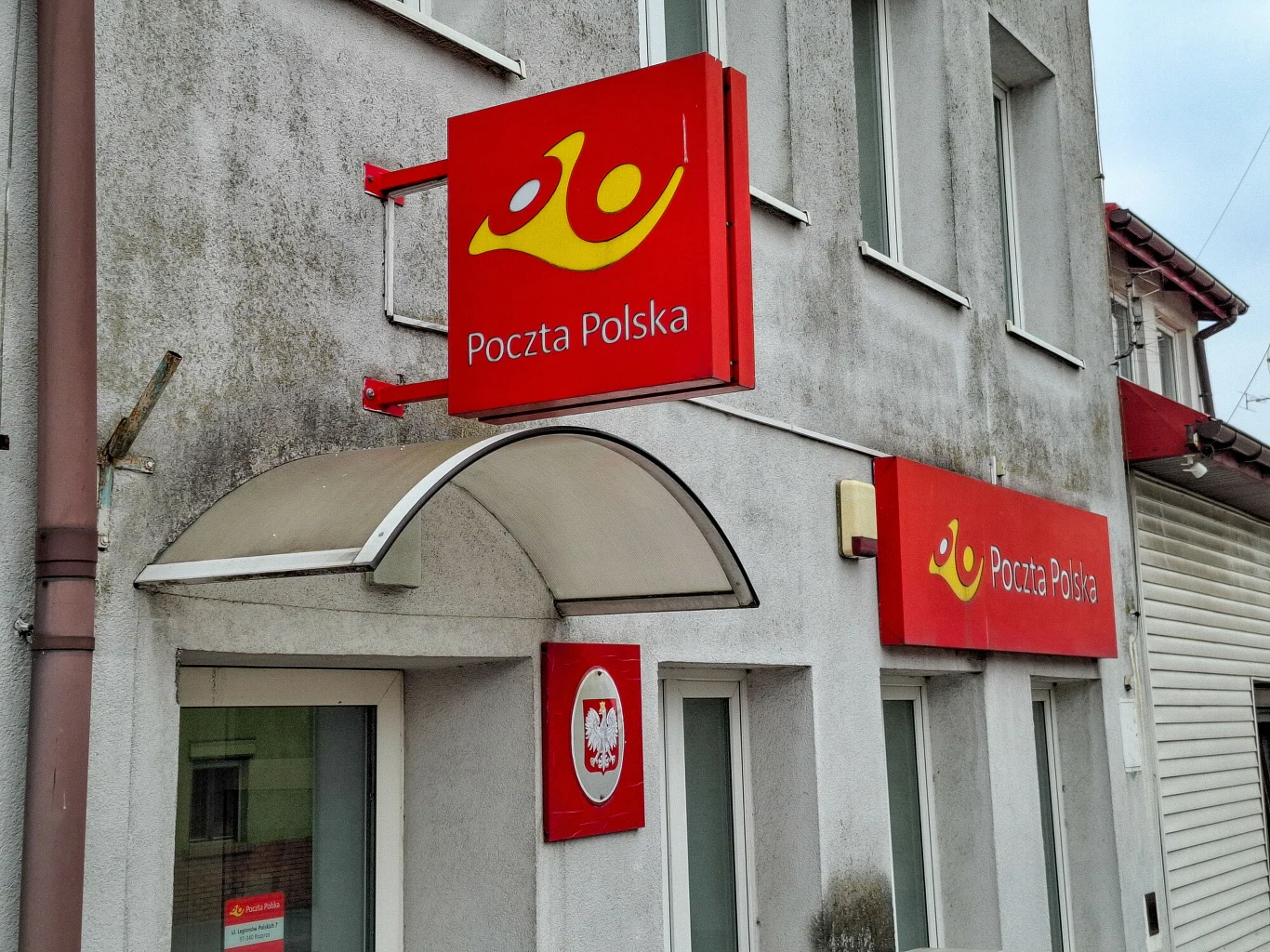 Poczta Polska zmienia zasady wysyłki, Polacy muszą przygotować się na zmiany (zdj. ilustracyjne) Fasada budynku z szyldami Poczty Polskiej, wejście osłonięte przezroczystym daszkiem, logo i napisy w barwach czerwono-żółtych, biała elewacja z widoczną tablicą z godłem Polski.