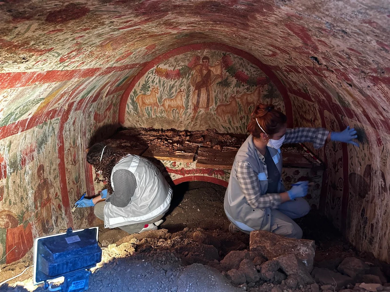 Dwóch archeologów w białych fartuchach, wyposażonych w maseczki ochronne i rękawice, prowadzi prace konserwatorskie w starożytnej krypcie ozdobionej kolorowymi freskami przedstawiającymi postacie i zwierzęta. Wewnątrz krypty widoczna jest ziemia oraz s...