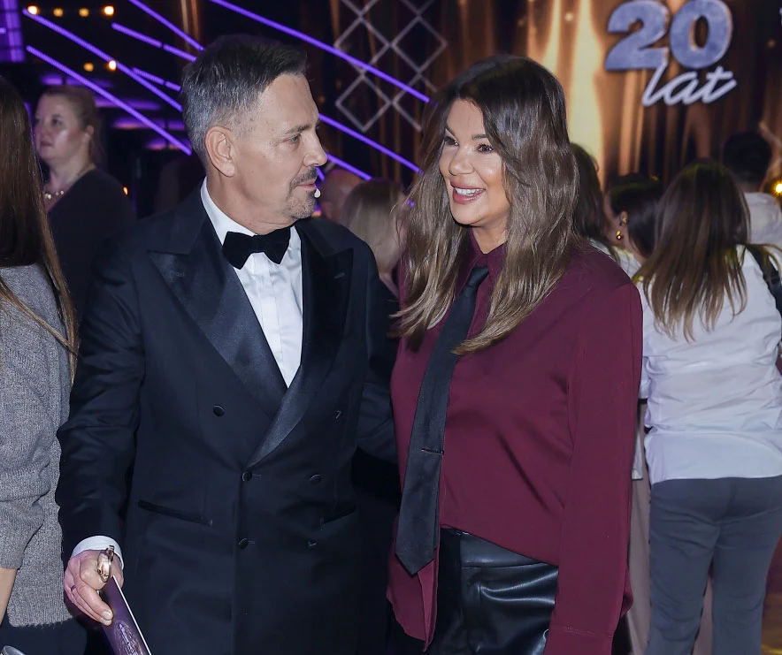 Krzysztof Ibisz i Edyta Górniak w "Tańcu z gwiazdami" Uśmiechnięta kobieta w bordowej koszuli i czarnej spódnicy oraz elegancko ubrany mężczyzna w smokingu stoją razem wśród innych osób w sali udekorowanej światłami i napisem 20 lat w tle. Panuje radosna, uroczysta atmosfera.