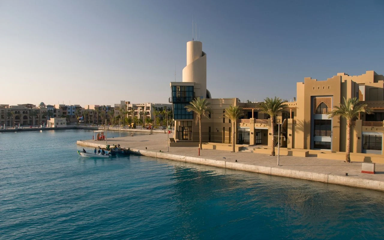 W Marsa Alam w dzielnicy Port Ghalib powstaje pierwsze "polskie osiedle" Nowoczesny budynek portowy z jasnymi elewacjami otoczony palmami przy spokojnej, błękitnej wodzie. W tle widoczne są kolejne budynki oraz łódź zacumowana przy betonowym nabrzeżu.