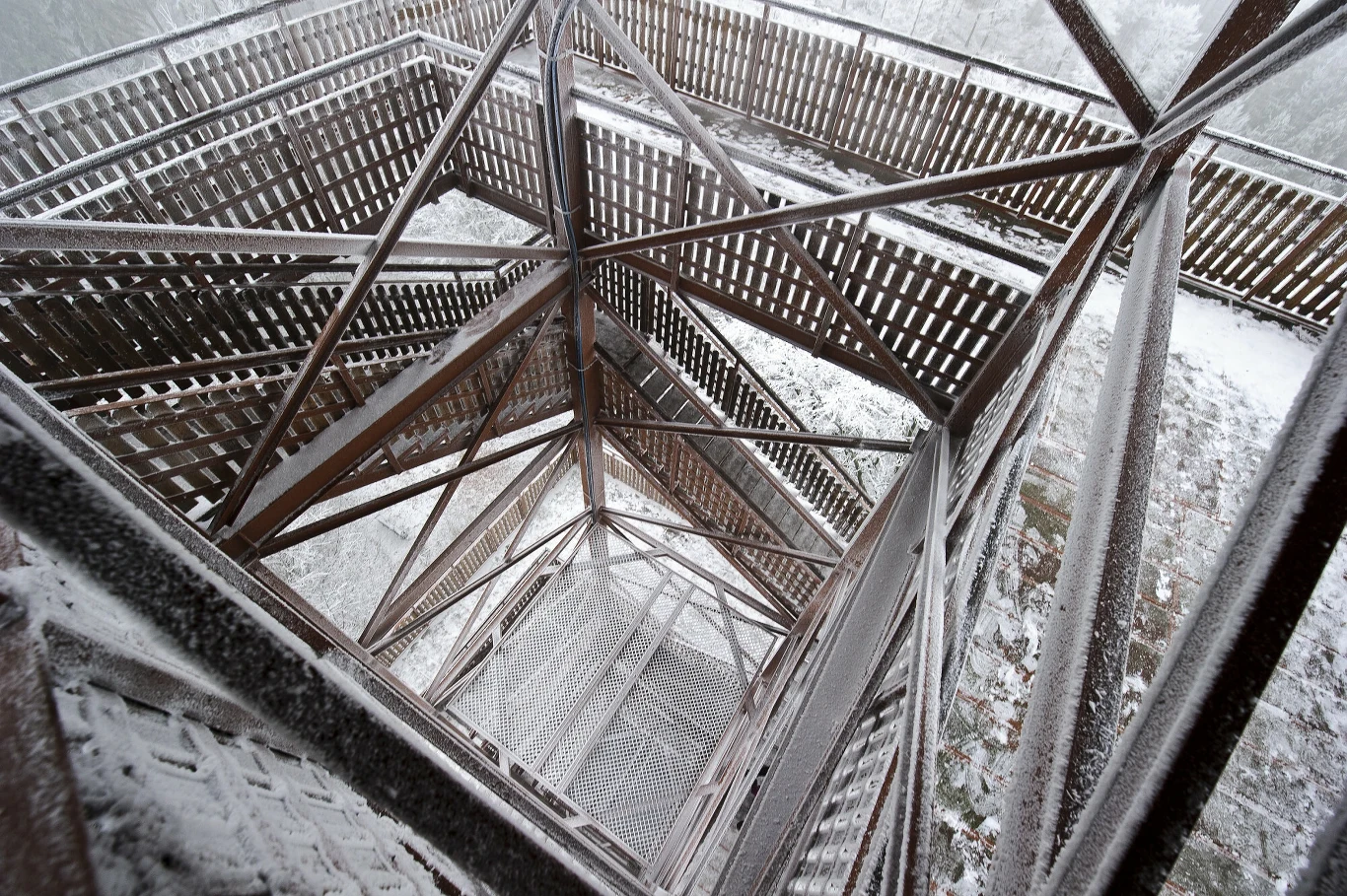 Metalowa konstrukcja wieży widokowej pokryta śniegiem, drewniane balustrady i geometryczny układ stalowych belek tworzą dynamiczną perspektywę skierowaną w dół.