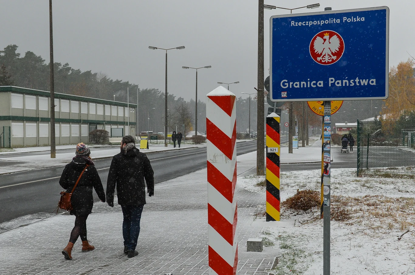 Boom w Polsce budzi niepokój w Niemczech. "Zostaliśmy w tyle" (zdj. ilustracyjne) Para ludzi idąca pieszo wzdłuż drogi przy przejściu granicznym, widoczne są słupki graniczne, znak informujący o granicy państwowej Polski oraz lekko zaśnieżone otoczenie.