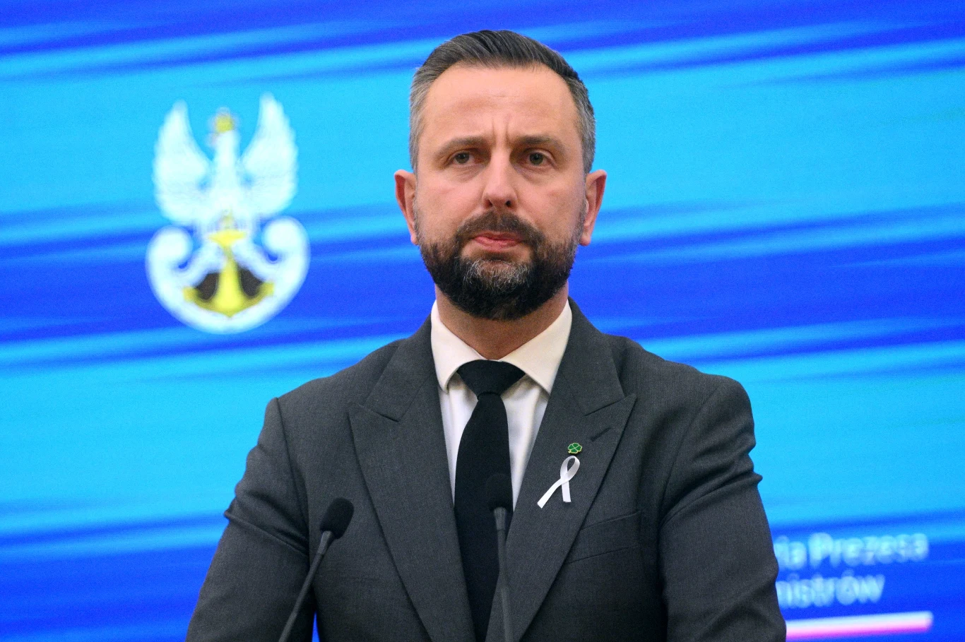 Polska. 39,1 proc. Polaków pozytywnie ocenia działania Władysława Kosiniaka-Kamysza w zakresie bezpieczeństwa i obronności kraju Mężczyzna w garniturze i pod krawatem stoi za mikrofonem na tle niebieskiego ekranu z orłem i wyraźnymi liniami graficznymi. Przypięta do klapy marynarki wstążka sugeruje udział w ważnym wydarzeniu lub kampanii społecznej.