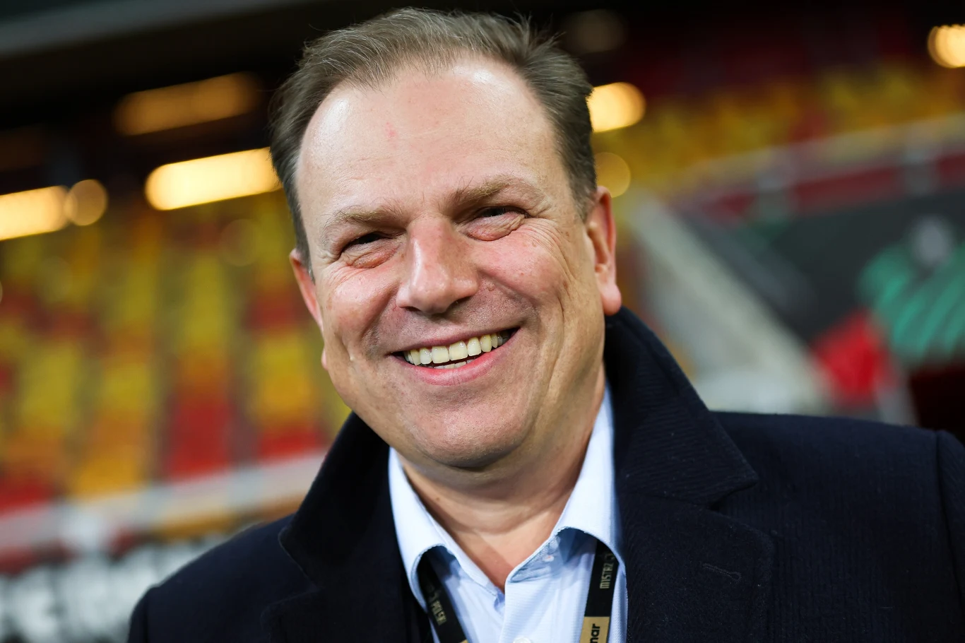 Zbliżenie na uśmiechniętego mężczyznę ubranego w ciemny płaszcz i jasną koszulę, stojącego na tle stadionu z miejscami dla widzów w kolorach czerwonym i żółtym.