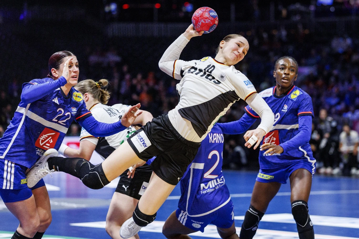 Zawodniczka w jasnym stroju piłki ręcznej oddaje rzut w kierunku bramki w trakcie dynamicznej akcji ofensywnej, podczas gdy dwie zawodniczki w niebieskich strojach próbują ją zablokować. Trybuny w tle podkreślają atmosferę sportowego wydarzenia.