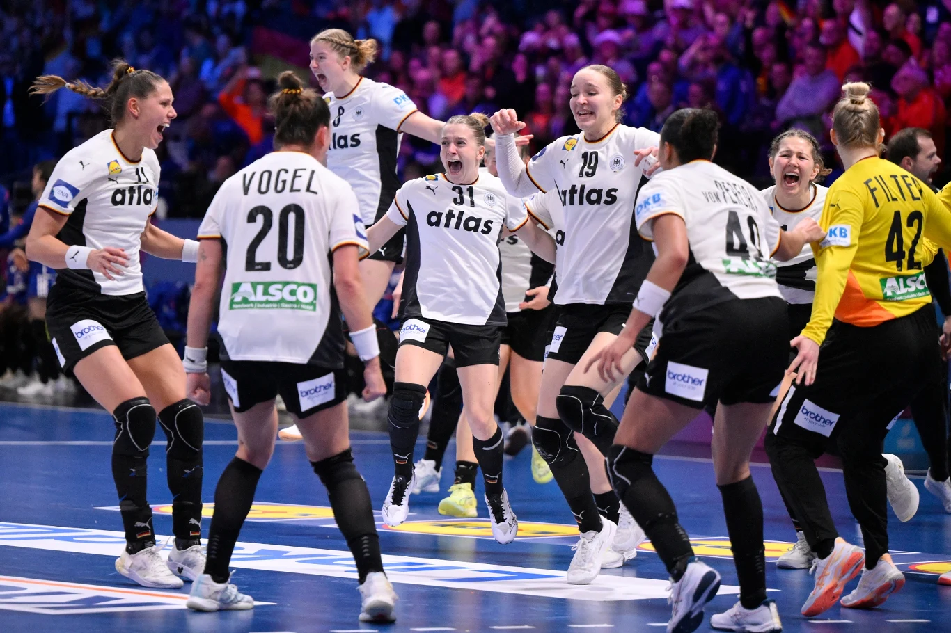 Zawodniczki drużyny piłki ręcznej w białych koszulkach świętują zwycięstwo na parkiecie, otoczone kibicami, pełne energii i radości, w tle widać trybuny oświetlone światłem reflektorów.