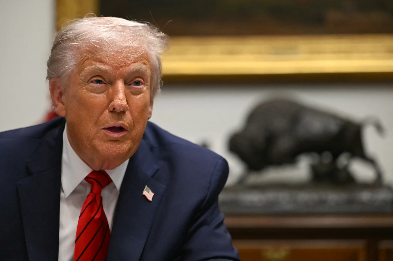 Prezydent USA Donald Trump Mężczyzna w garniturze z czerwonym krawatem siedzący przy stole, w tle widoczna rzeźba byka oraz fragment obrazu w złotej ramie.