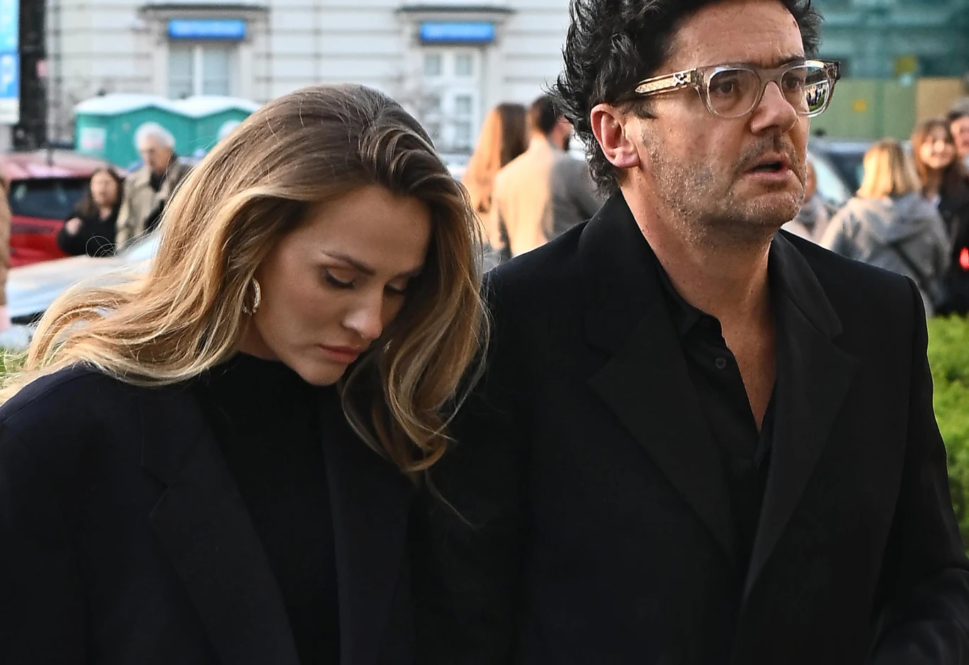 Kuba Wojewódzki z Żanetą Kobieta i mężczyzna idą razem, oboje ubrani elegancko w ciemne ubrania, kobieta ma spuszczoną głowę, mężczyzna patrzy przed siebie. W tle widoczne są rozmazane sylwetki ludzi oraz elementy miejskiej architektury.