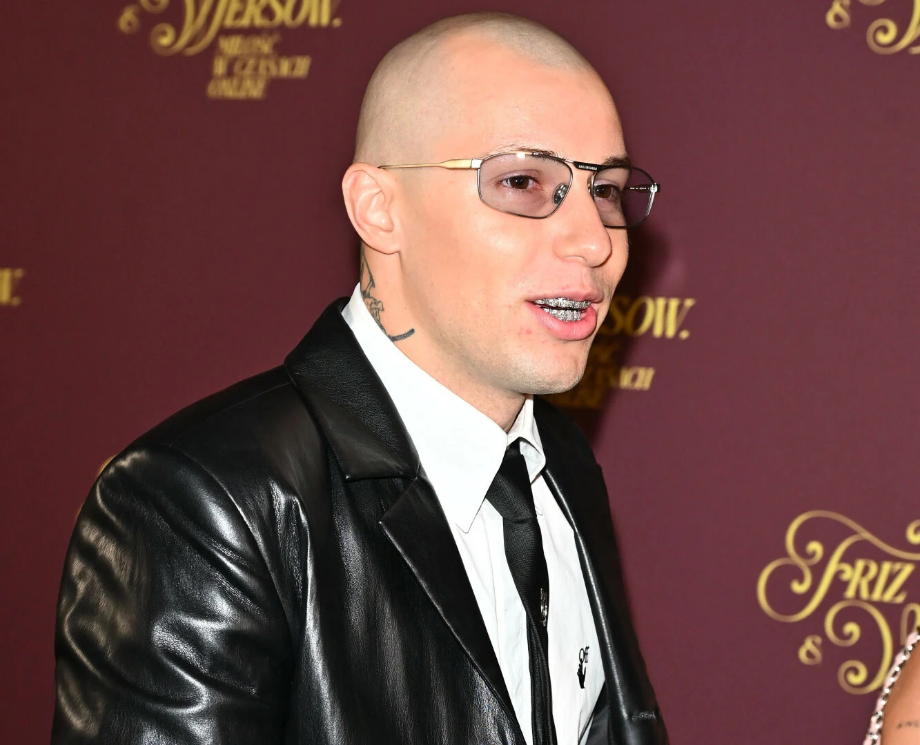 Żabson nagrał duet z Wizem Khalifą Mężczyzna z ogoloną głową, w okularach przeciwsłonecznych, ubrany w czarną skórzaną kurtkę, białą koszulę oraz czarny krawat, pozuje na tle bordowej ścianki z złotymi napisami.