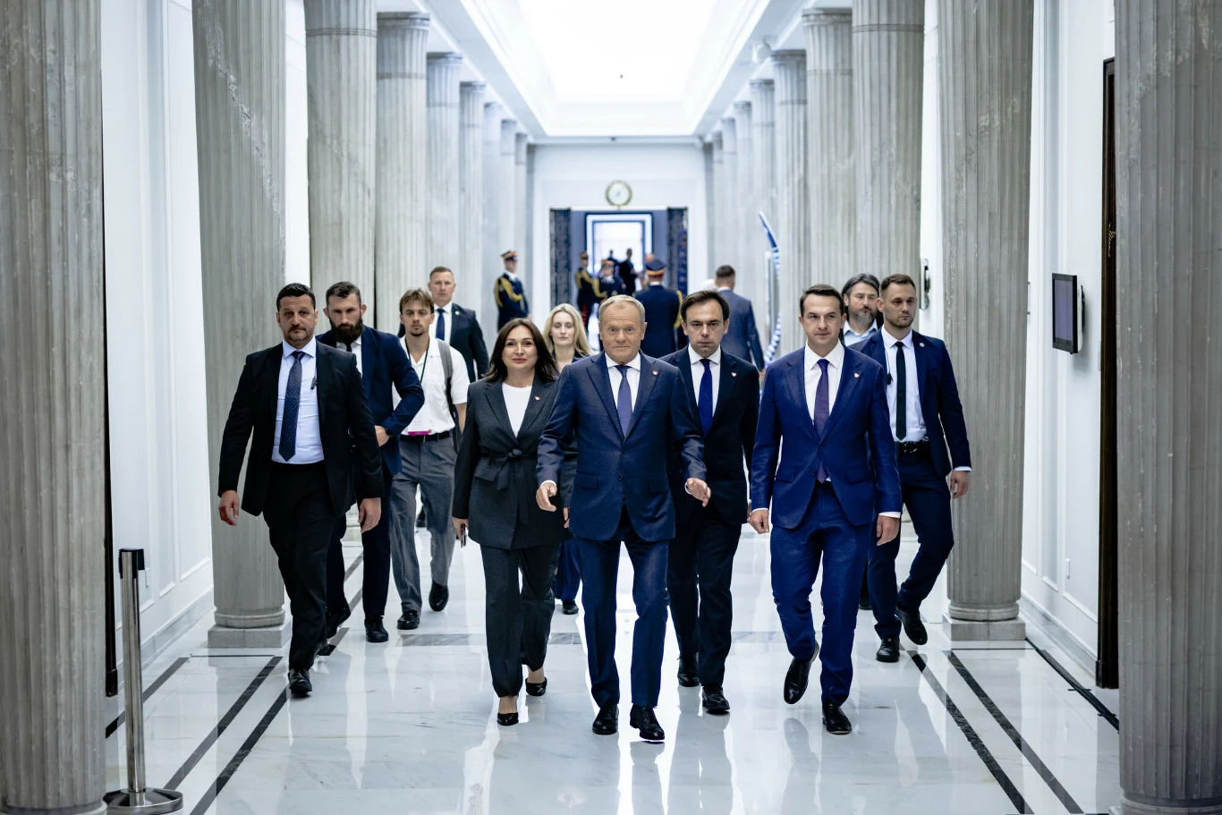 Rząd Donalda Tuska sprawuje władzę od 13 grudnia 2023 roku Grupa elegancko ubranych osób idzie korytarzem w budynku rządowym, przewodzi im mężczyzna w garniturze, po obu stronach towarzyszą mu inni politycy, w tle widać kolumny oraz drzwi prowadzące do kolejnych pomieszczeń.