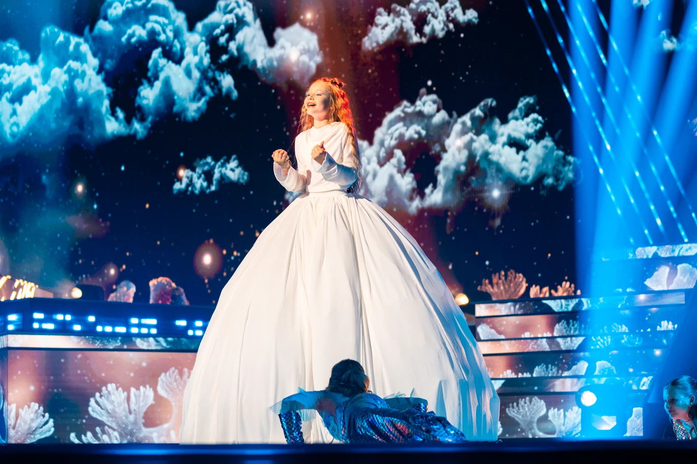 Marianna Kłos reprezentuje Polskę podczas Eurowizji Junior 2025 Artystka w obszernej białej sukni stoi na scenie oświetlonej jasnymi światłami, w tle dynamiczna scenografia z chmurami i błękitnym niebem oraz aktor leżący u jej stóp w połyskującym stroju.