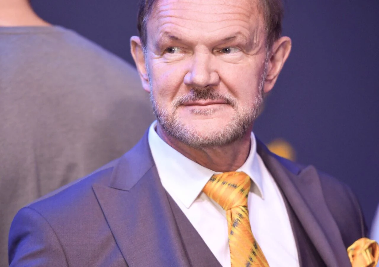 Cezary Pazura Mężczyzna w średnim wieku w eleganckim garniturze z białą koszulą i żółtym krawatem, lekko uśmiechnięty, patrzy w bok. W tle rozmyta postać i niebieskie tło.