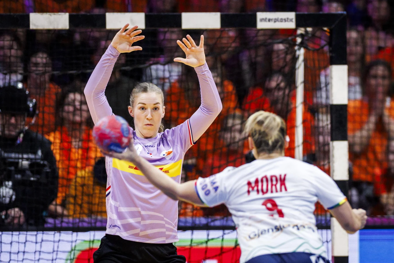 Zawodniczka piłki ręcznej w bramce podnosi ręce do obrony, naprzeciwko niej przeciwniczka przygotowuje się do rzutu piłką, a w tle widownia wypełniona kibicami.