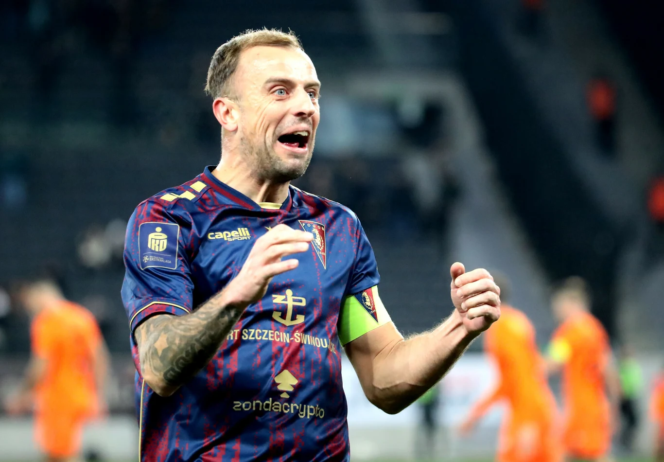 Piłkarz w granatowo-bordowym stroju z opaską kapitana na ramieniu, wyrażający silne emocje na tle rozmytych zawodników ubranych w pomarańczowe stroje na boisku sportowym.