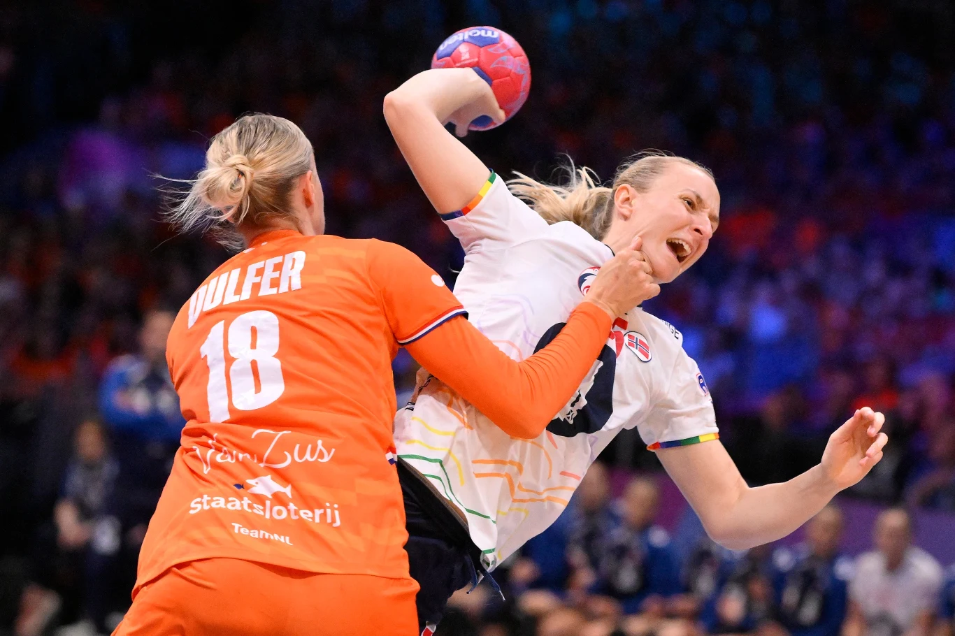 Intensywna walka podczas meczu piłki ręcznej kobiet, zawodniczka w białej koszulce rzuca piłkę, jednocześnie będąc blokowaną przez przeciwniczkę w pomarańczowym stroju. W tle rozmazana publiczność i kolorowe światła hali sportowej.