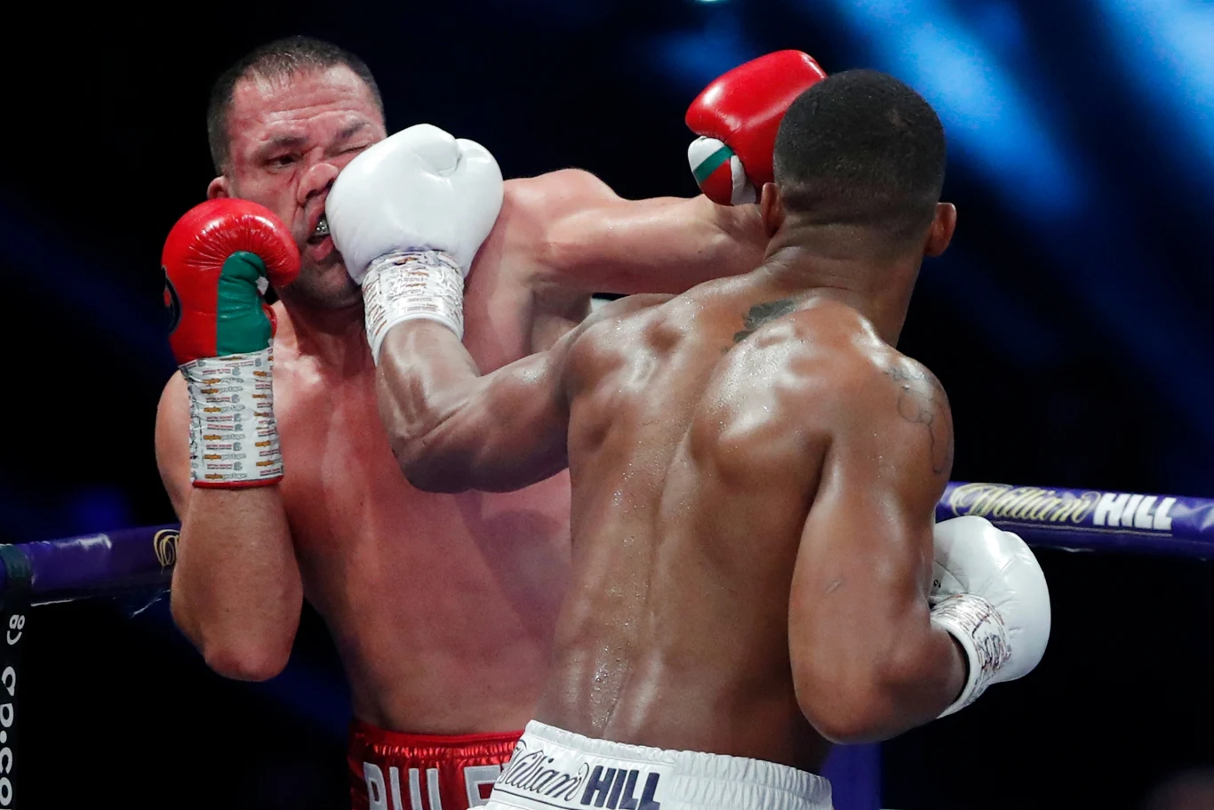 Kubrat Pulew (z lewej) Dwu bokserów w ringu, jeden z nich otrzymuje silny cios w twarz, co podkreśla dynamikę i intensywność walki. Sportowa atmosfera, rękawice bokserskie i bandy widoczne w tle, skupienie na akcji podczas pojedynku.