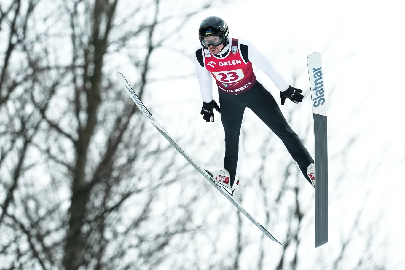 Witalij Kaliniczenko Sportowiec w trakcie skoku narciarskiego, ubrany w strój sportowy, kask oraz gogle, unosi się w powietrzu z rozpostartymi nartami, w tle rozmyte drzewa.