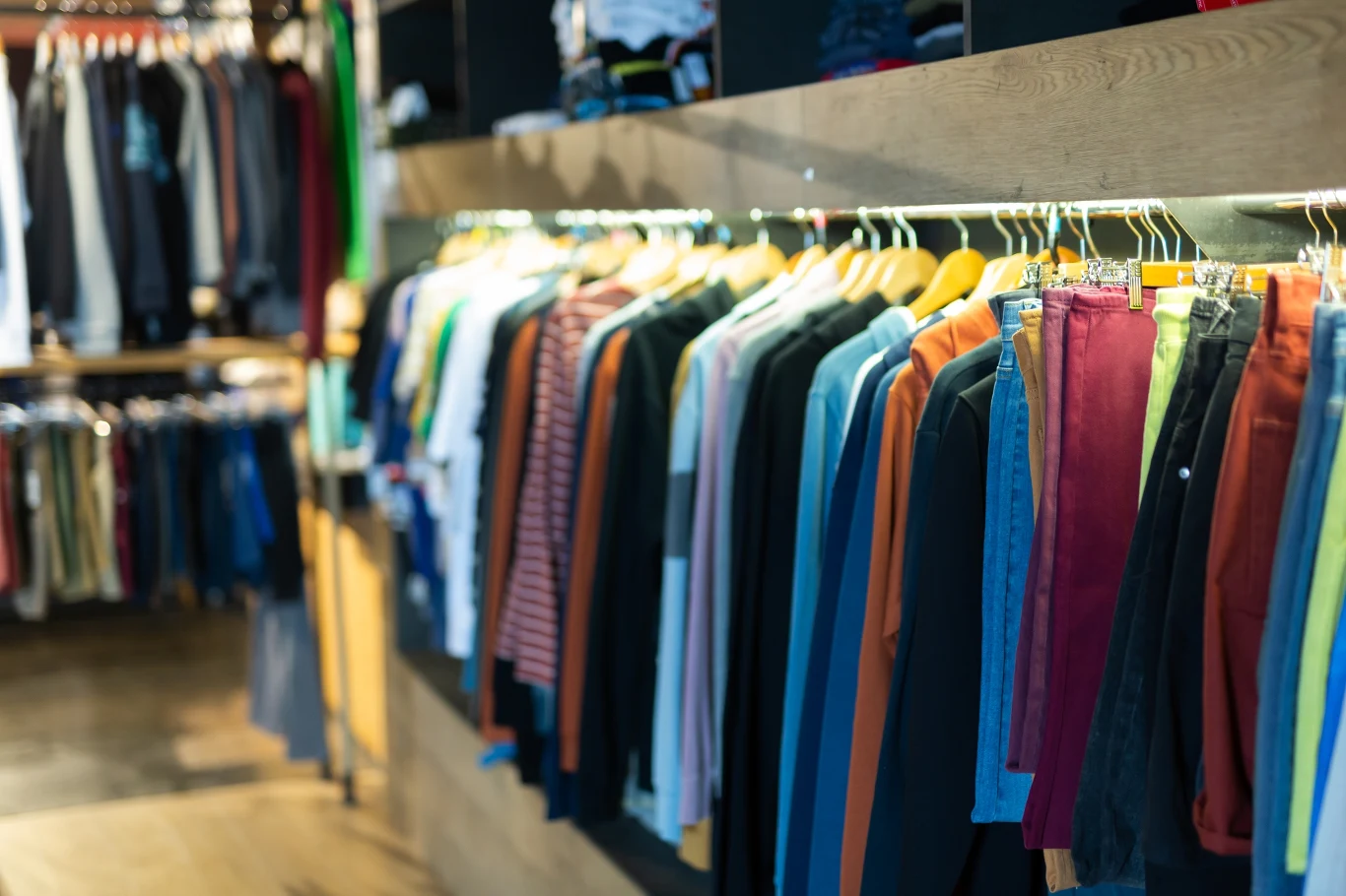 Uważaj co wkładasz. Ubrania mogą być także toksyczne Rząd kolorowych koszul i bluzek wiszących na wieszakach w nowoczesnym sklepie odzieżowym, podświetlonych światłem LED, z widoczną częścią drewnianego regału i drewnianą podłogą.