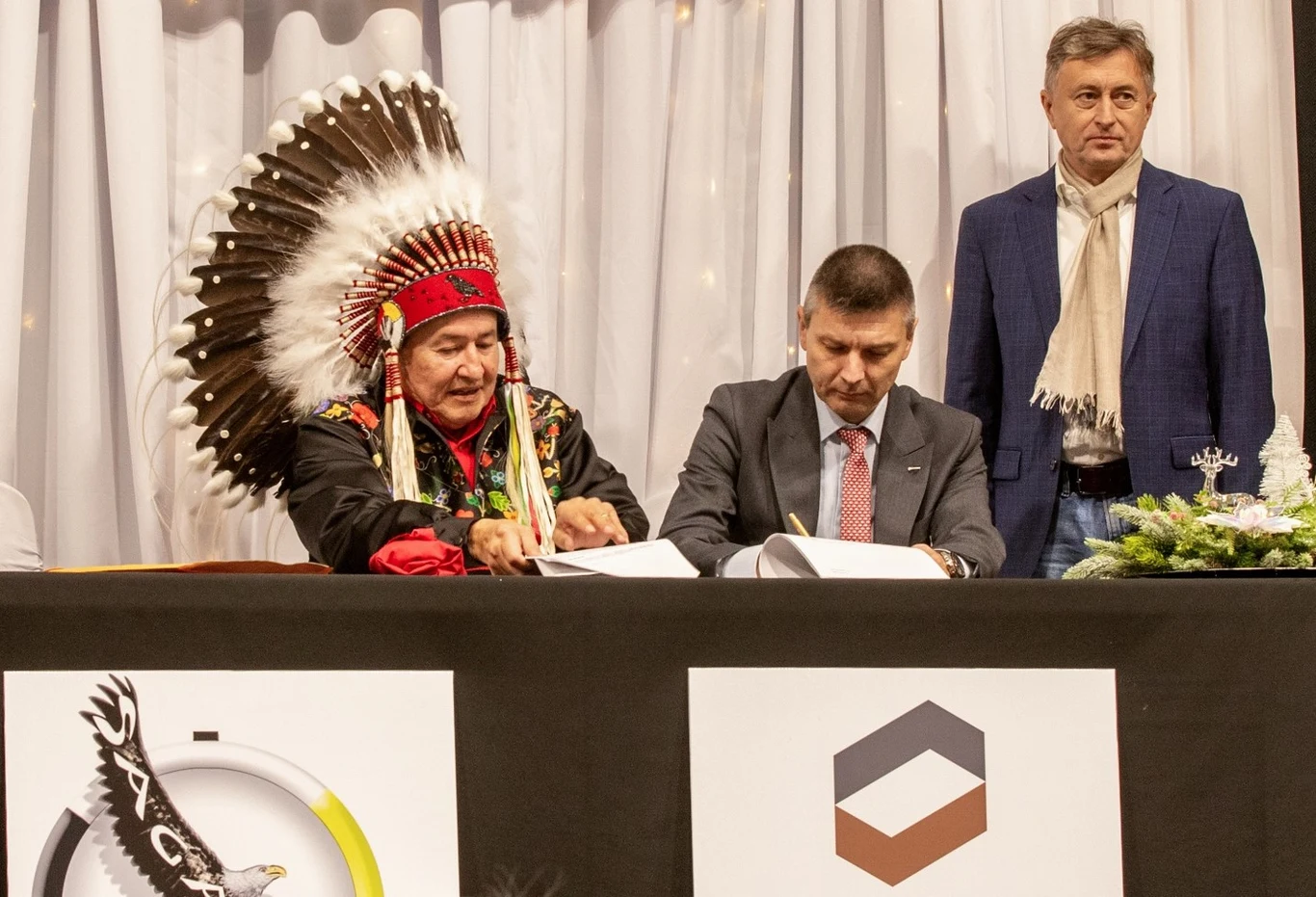 KGHM zawarł umowę z rdzenną ludnością w Kanadzie Osoba w tradycyjnym pióropuszu indiańskim oraz dwóch mężczyzn w garniturach siedzą przy stole i podpisują dokumenty, w tle biała zasłona i świąteczna dekoracja, na stole dwa logotypy.