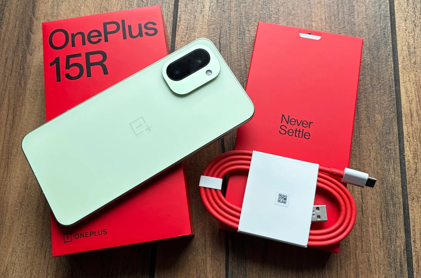 Nowy smartfon OnePlus 15R w jasnej obudowie, czerwony kabel USB-C oraz czerwona oryginalna ładowarka, wszystko na drewnianym tle. Obok urządzenia widoczne kartonowe opakowanie z wygrawerowaną nazwą modelu.