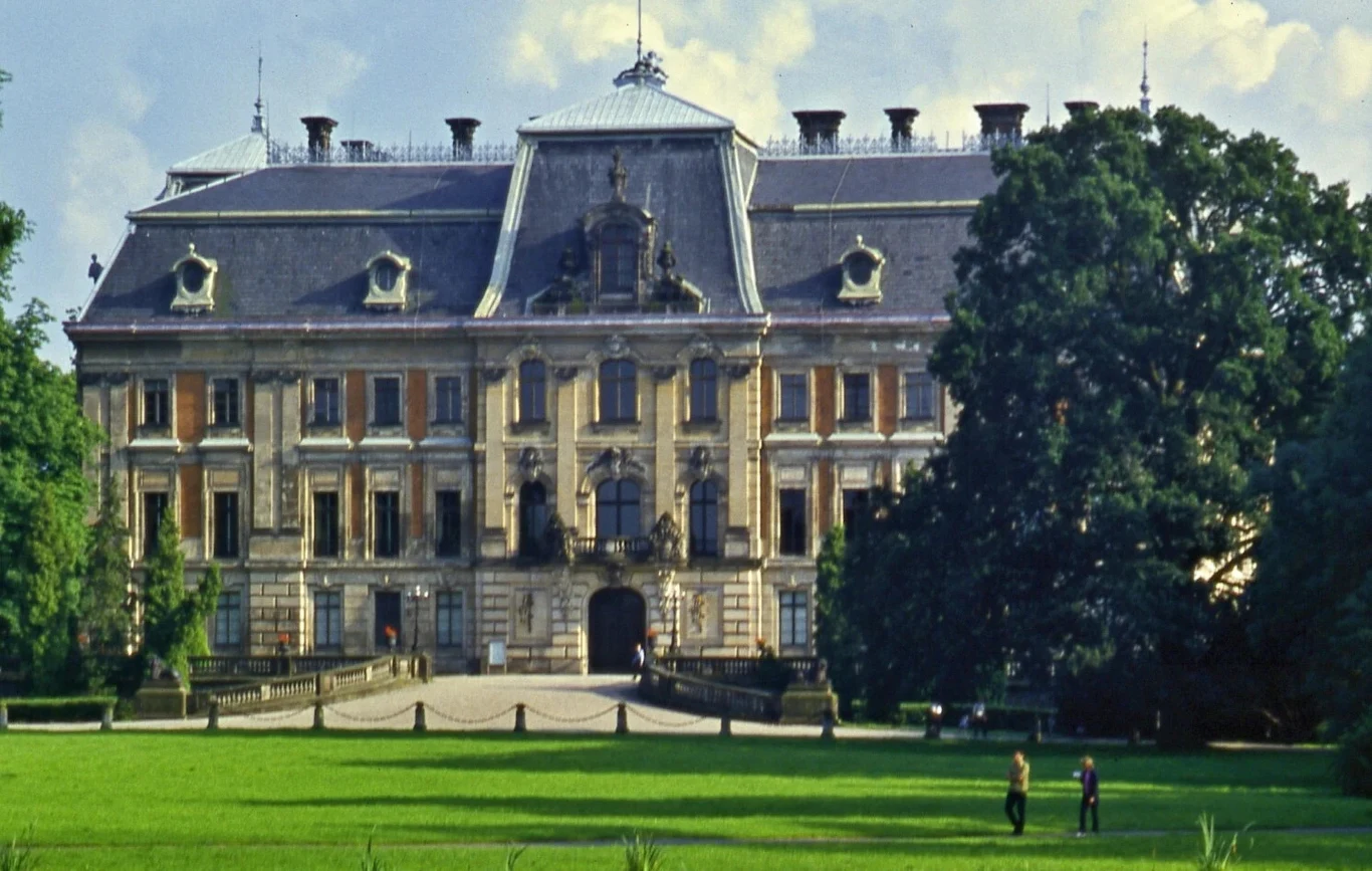 Pszczyna szczyci się pięknym zespołem pałacowo-parkowym. Czemu mówimy o Pszczynie a nie "Przczynie"? Okazały pałac z wysokim dachem mansardowym i bogato zdobioną elewacją frontową, otoczony zielenią oraz dużą trawiastą przestrzenią, na której widoczne są dwie osoby spacerujące w pobliżu okazałego drzewa.