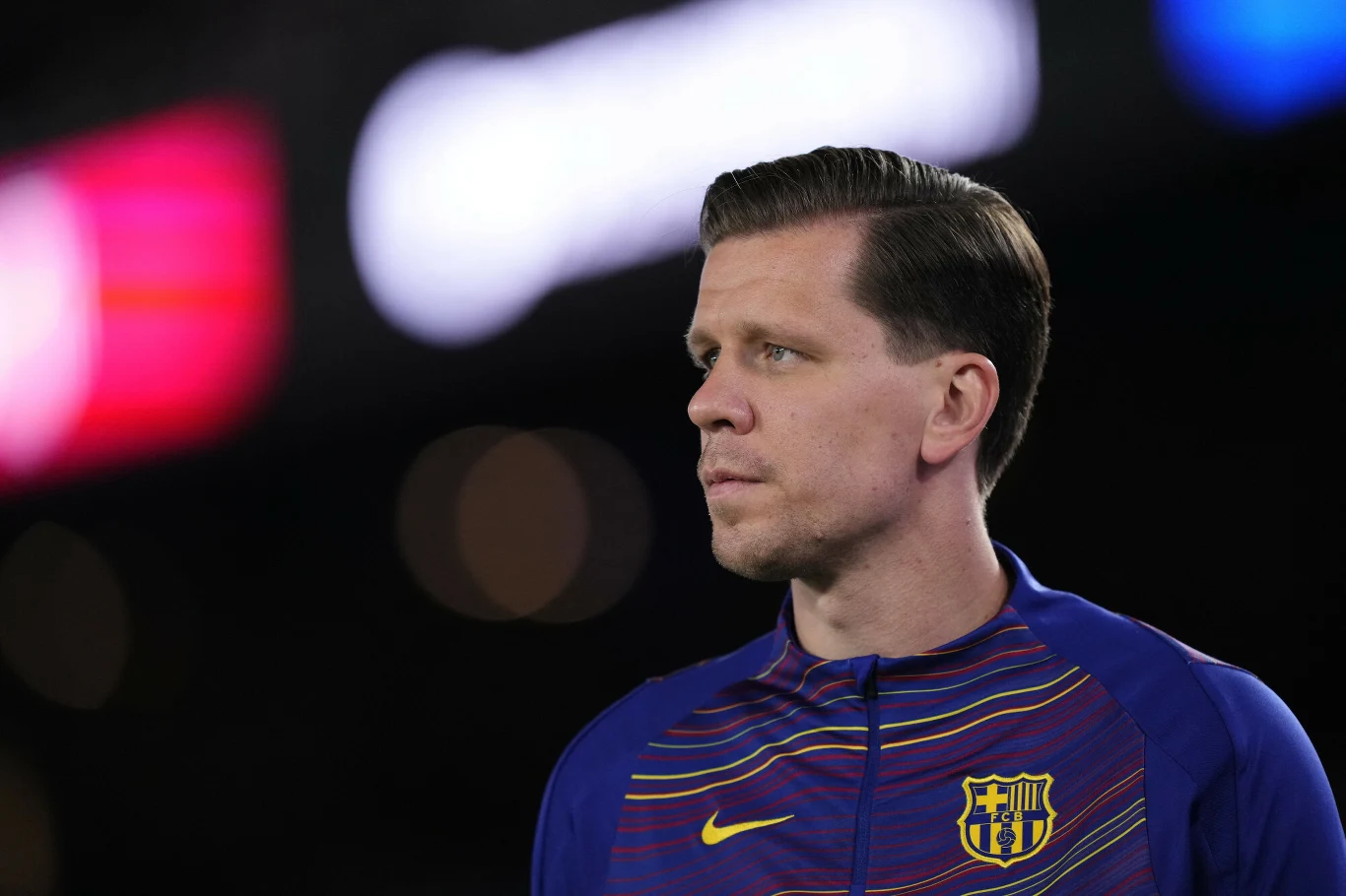 Wojciech Szczęsny Mężczyzna w sportowej bluzie z emblematem FC Barcelony patrzy w bok w kierunku jasnych świateł na rozmytym tle.