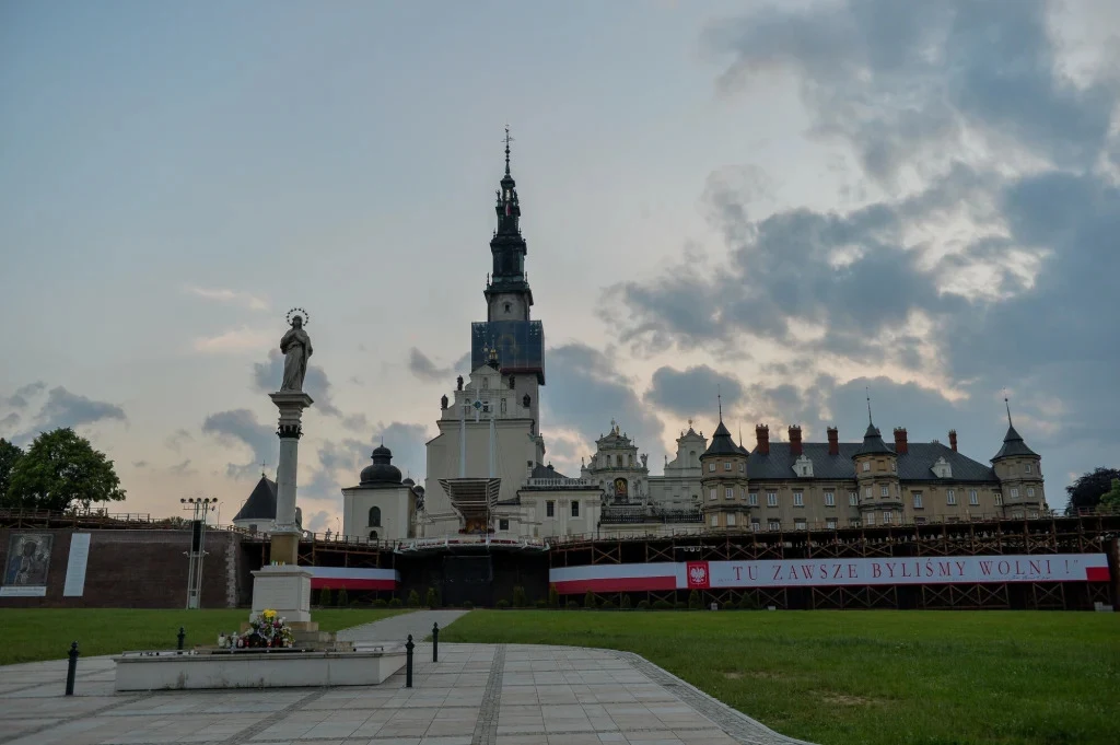 Według legend Częstochowa miała się chować za wzgórzami przed napływającymi pielgrzymami Widok na Sanktuarium na Jasnej Górze w Częstochowie, z wyraźnie widoczną wieżą klasztoru, otaczającymi zabudowaniami oraz pomnikiem Matki Bożej na pierwszym planie. Na dole widoczny trawnik i chodnik, na tle zachodzącego słońca i pochmurnego nieba. W t...