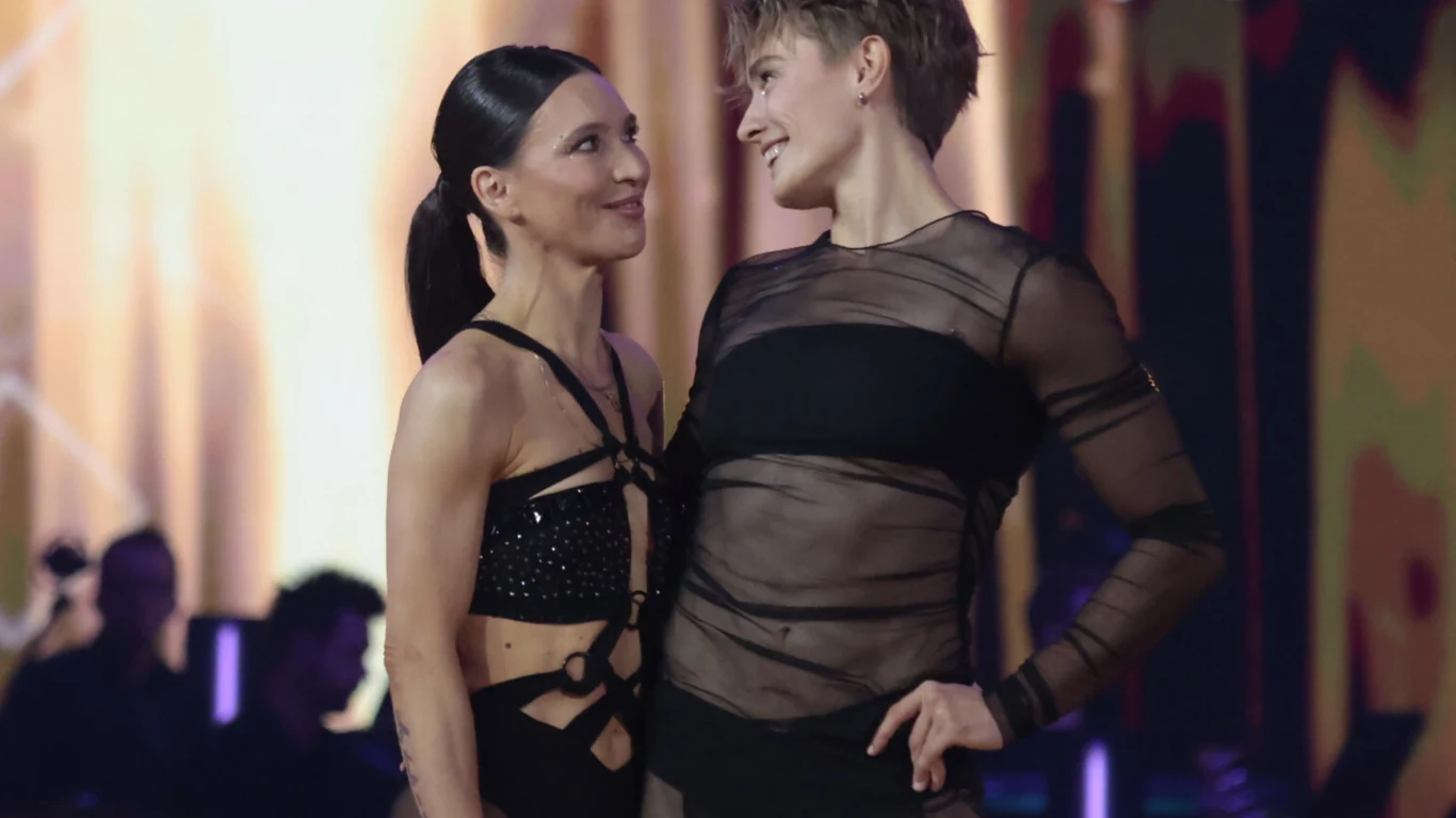 Dwie osoby w stylizowanych, tanecznych strojach stoją naprzeciwko siebie na scenie z kolorowym, rozświetlonym tłem. Oboje uśmiechają się i patrzą na siebie z wyraźnym zrozumieniem oraz ciepłymi emocjami.