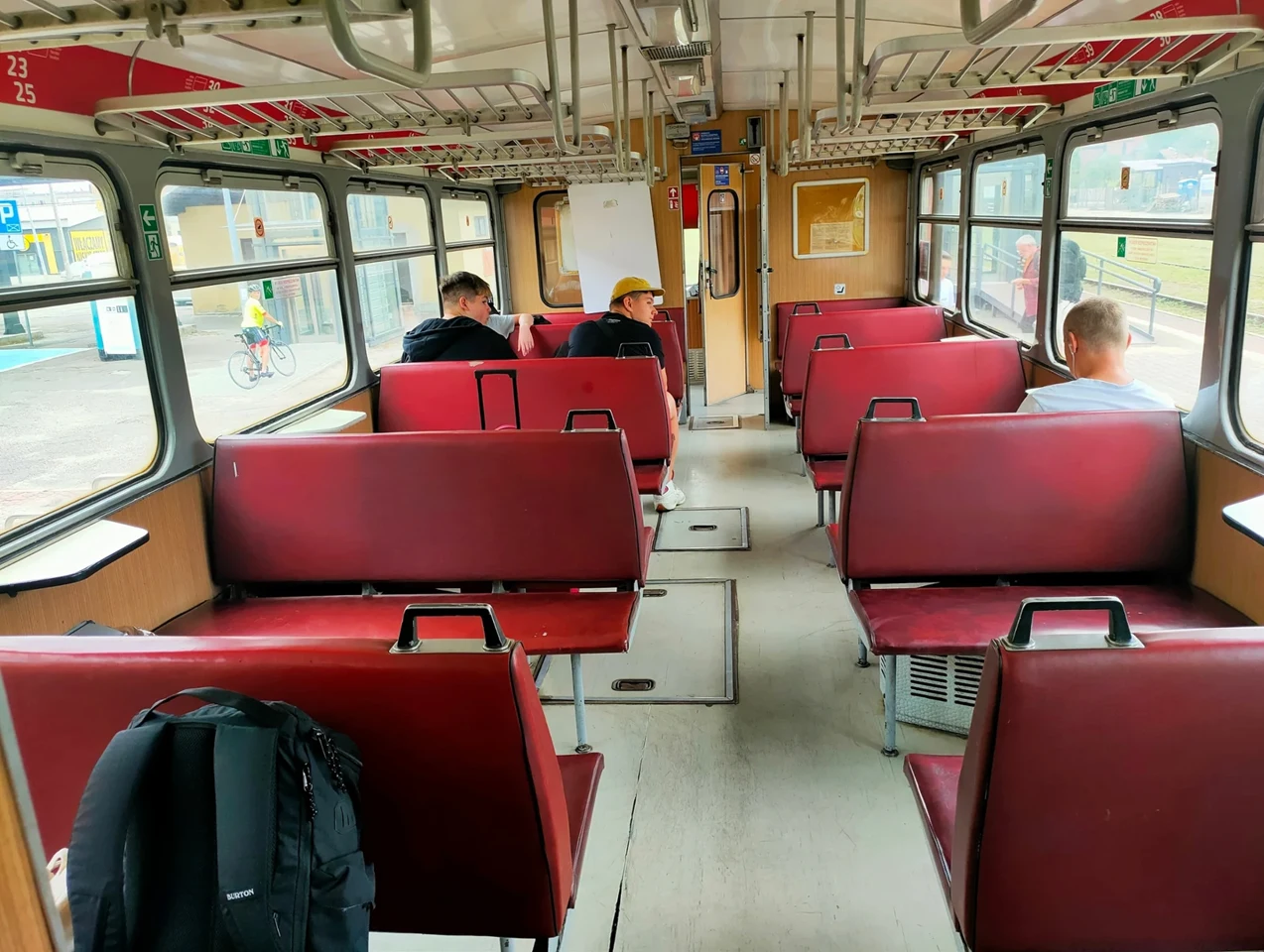 Wnętrze wagonu pociągu z czerwonymi siedzeniami, trzema pasażerami siedzącymi po przeciwnych stronach wagonu oraz plecakiem pozostawionym na pierwszym siedzeniu. Przez okna widać fragment peronu i przechodzącą osobę na rowerze.