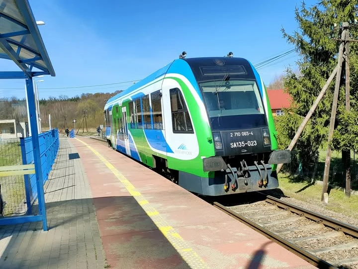 Na niektórych podkarpackich liniach pociągów jest sporo. Gdzie indziej oferta bywa dość skromna Nowoczesny, niebiesko-zielony pociąg zatrzymany na niewielkiej stacji kolejowej, otoczony niewysokim ogrodzeniem, z widoczną infrastrukturą peronową i fragmentami zabudowań pośród drzew w tle.