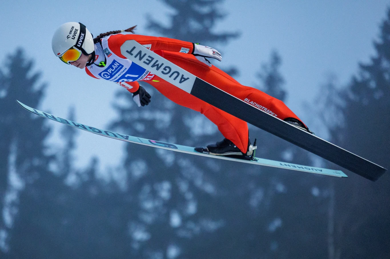 Skoczek narciarski w czerwonym kombinezonie podczas lotu, dynamicznie unosi się nad rozmytym tłem z sylwetkami drzew, wyposażony w kask oraz gogle, trzyma pozycję lotną na nartach z logo sponsora.