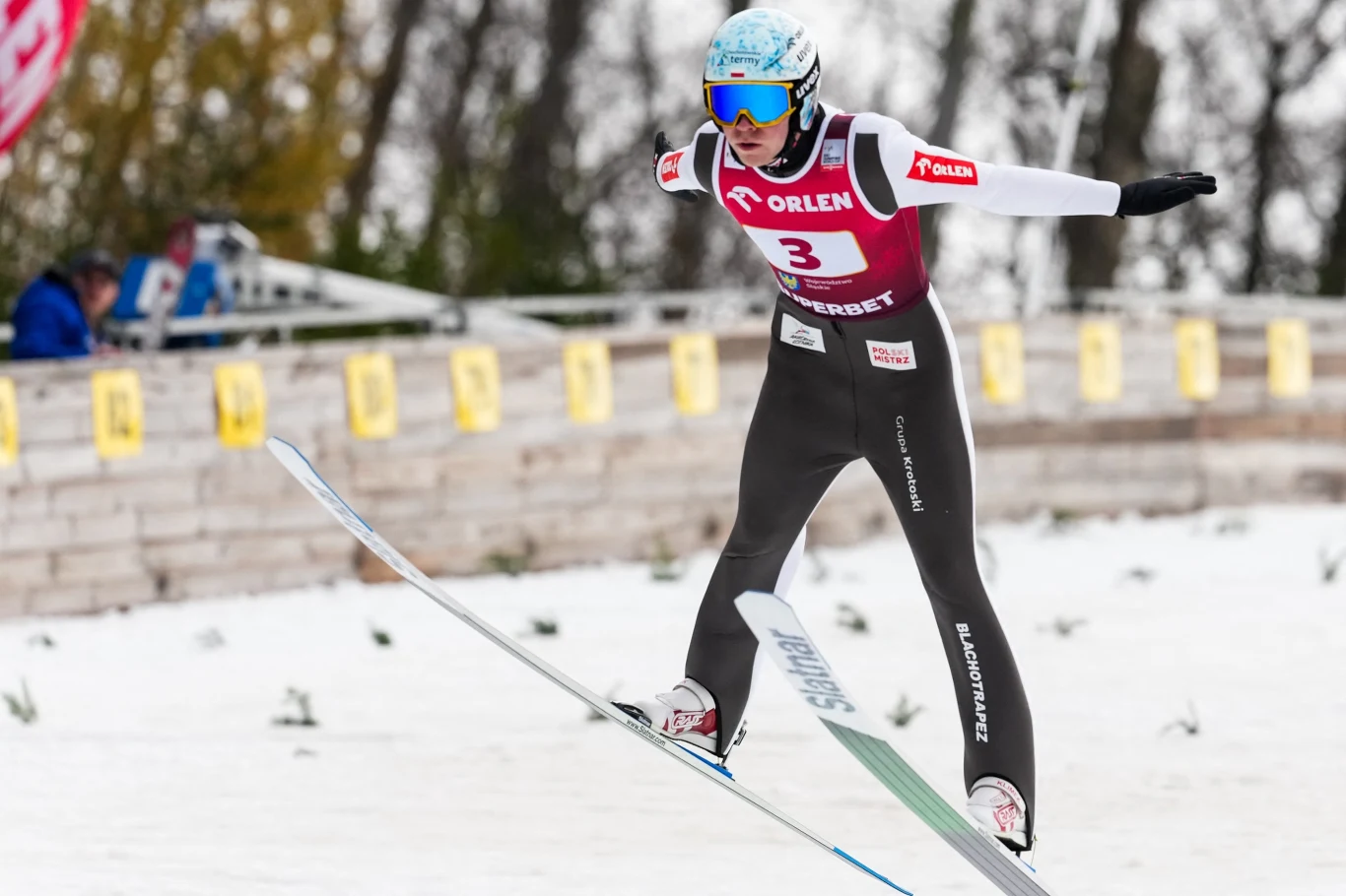 Zawodnik w trakcie skoku narciarskiego ubrany w kombinezon sportowy i kask, z uniesionymi ramionami, lecący nad zaśnieżonym podłożem, w tle rozmazane sylwetki drzew oraz tablice reklamowe.