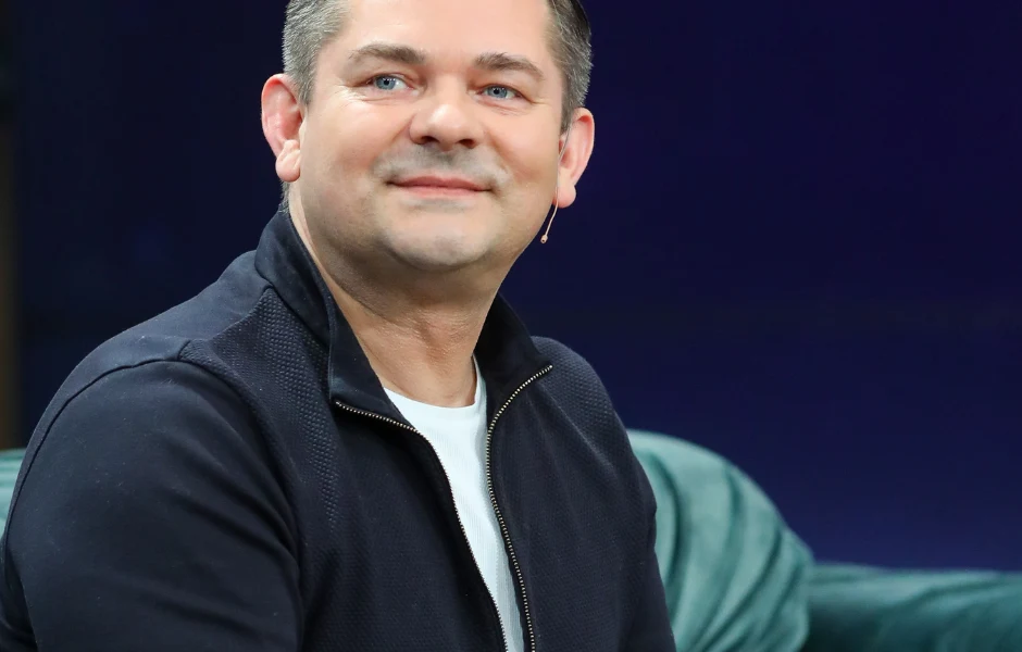 Zenon Martyniuk Uśmiechnięty mężczyzna ubrany w ciemną kurtkę i biały t-shirt siedzi na zielonej kanapie, w tle jednolite ciemne tło.