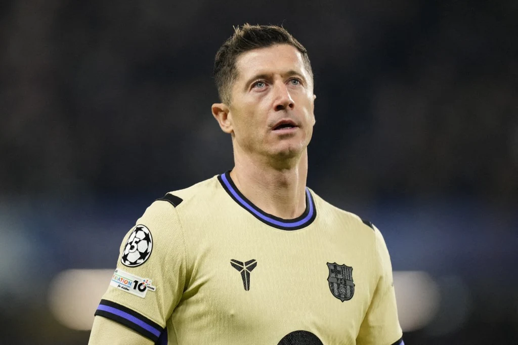 Piłkarz w kremowym stroju drużyny FC Barcelona patrzący w górę, w tle rozmyte trybuny stadionu. Na koszulce widoczne są naszywki Ligi Mistrzów oraz logo klubu.