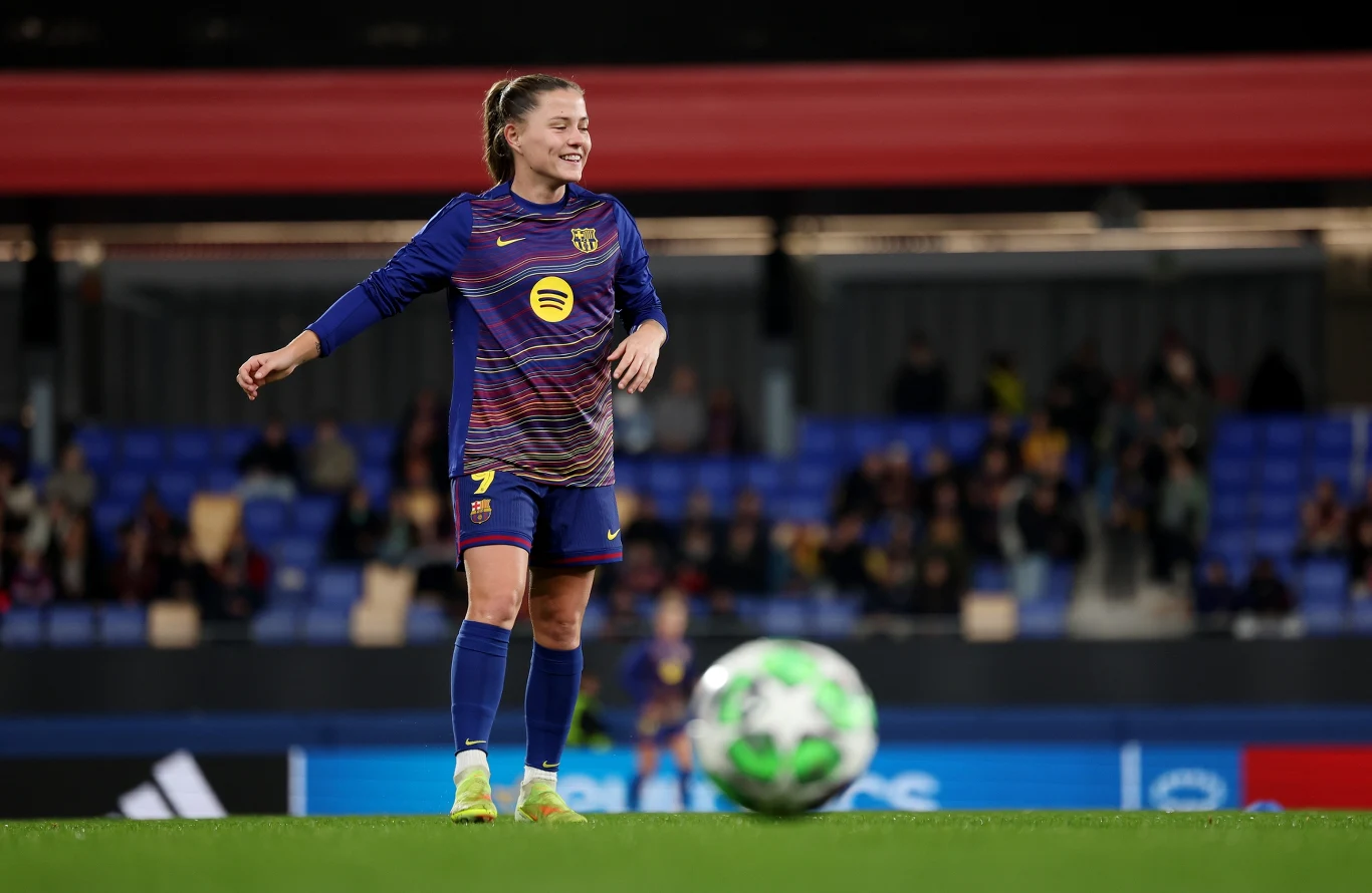 Claudia Pina Piłkarka w stroju FC Barcelony stoi na murawie stadionu z uśmiechem na twarzy, przygotowana do gry, w tle widoczna publiczność na trybunach.