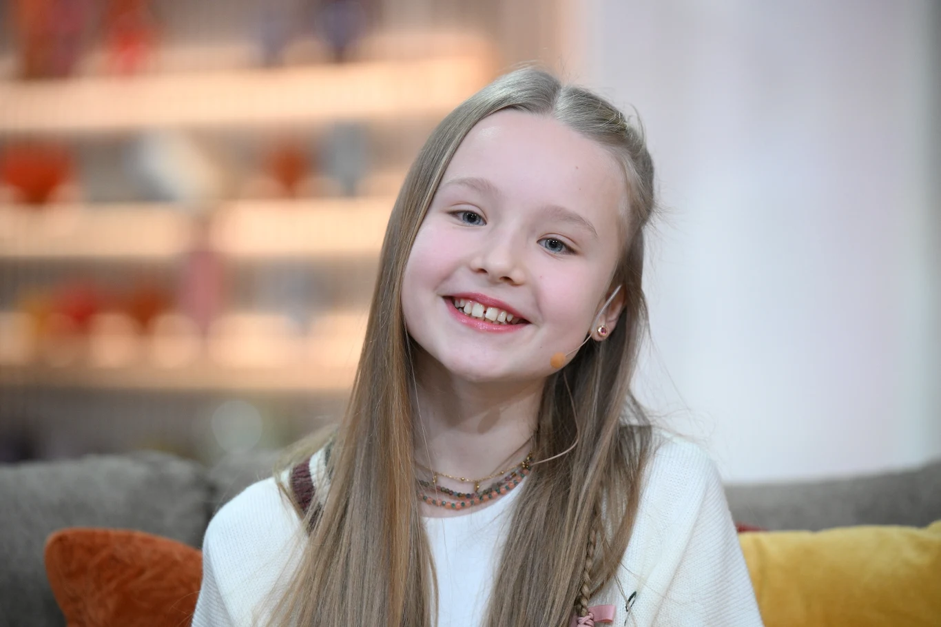 Marianna Kłos Uśmiechnięta dziewczynka z długimi blond włosami siedzi na sofie w jasnym pomieszczeniu, w tle rozmyte półki z kolorowymi elementami.