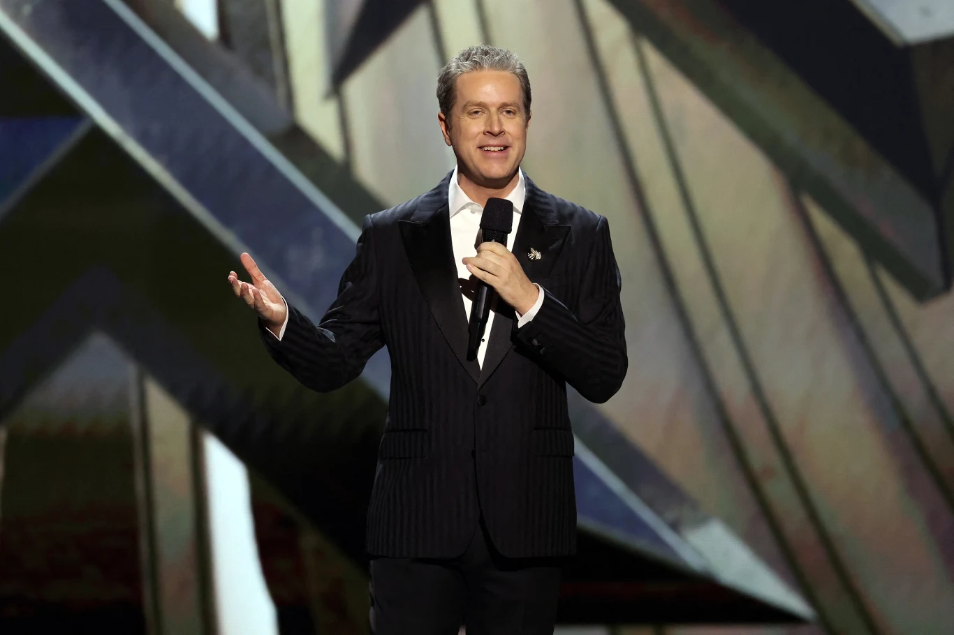 Geoff Keighley, prowadzący galę The Game Awards Mężczyzna w garniturze z mikrofonem w dłoni stoi na scenie, w tle widoczne geometryczne wzory w stonowanych barwach.