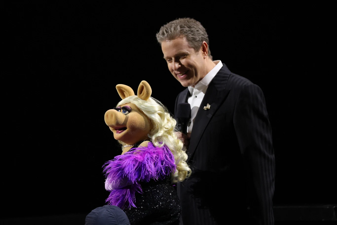 The Game Awards Mężczyzna ubrany w ciemny garnitur trzyma mikrofon i występuje na scenie obok znanej lalki świnki z blond peruką i ubraniem w fioletowym kolorze.